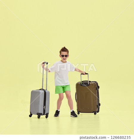 Smiling boy in white t-shirt and green shorts standing with two travel suitcases, posing confidently in sunglasses against yellow studio background Smiling boy in white t-shirt and green shorts standing with two travel suitcases, posing confidently in sunglasses against yellow studio background 126680235