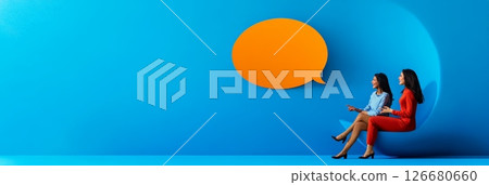 Two businesswomen sitting on a crescent moon, discussing under a large orange speech bubble against a vibrant blue backdrop, symbolize modern communication and innovative thinking 126680660