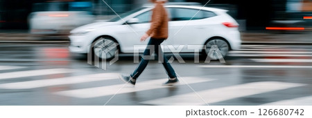 Young man walking on a pedestrian crossing while a white car speeds by on a wet city street, creating a near miss and highlighting the dangers of urban traffic 126680742