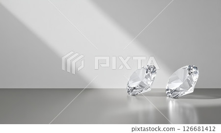 Two brilliant cut diamonds rest on a gray surface, with a white wall in the background and soft, directional light creating highlights and shadows, providing a luxurious and elegant backdrop 126681412