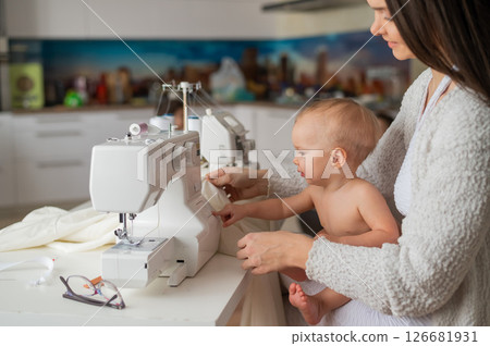 A young woman sews in the kitchen and holds a small child. Mom teaches her little son to sew on a sewing machine. Self-isolation homework A young woman sews in the kitchen and holds a small child. Mom teaches her little son to sew on a sewing machine. Self-isolation homework 126681931