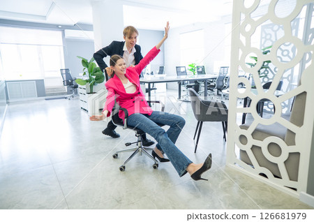 Coworkers fooling around in the office. A guy pushes a young woman around on a chair.  126681979