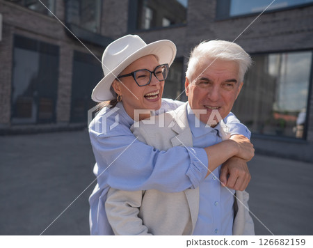 A woman in a hat and glasses hugs an elderly man in a white jacket from behind. Romantic relationships of mature people. A woman in a hat and glasses hugs an elderly man in a white jacket from behind. Romantic relationships of mature people. 126682159