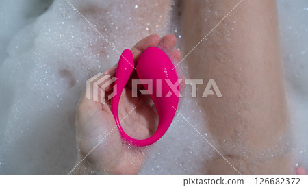 A woman lies in a bubble bath and holds a curved pink vibrator. A woman lies in a bubble bath and holds a curved pink vibrator. 126682372