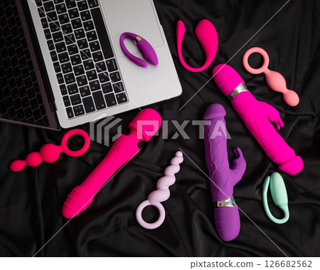 Collection of sex toys on a black sheet. Top view of various vibrators and a laptop.  126682562
