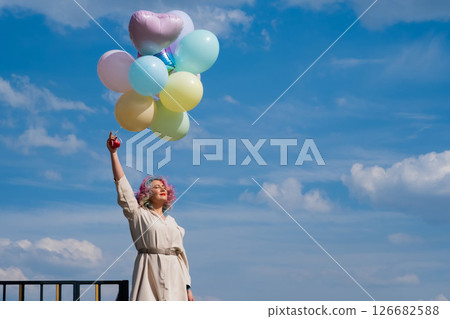 A woman with colored hair with an armful of balloons against a blue sky. A woman with colored hair with an armful of balloons against a blue sky. 126682588