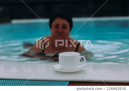 A white cup and sunglasses sit beside a pool with a person enjoying a swim on a sunny day. A relaxing scene and a person in the pool, summer leisure. A white cup and sunglasses sit beside a pool with a person enjoying a swim on a sunny day. A relaxing scene and a person in the pool, summer leisure. 126682638
