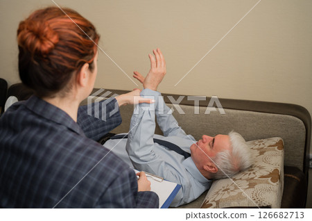 An elderly man lies on a couch and holds his hands hanging during an appointment with a hypnotherapist. An elderly male patient in an individual consultation with a female psychologist An elderly man lies on a couch and holds his hands hanging during an appointment with a hypnotherapist. An elderly male patient in an individual consultation with a female psychologist 126682713