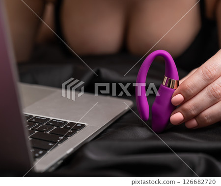 A woman is lying in bed holding a curved vibrator and looking at a laptop. A woman is lying in bed holding a curved vibrator and looking at a laptop. 126682720