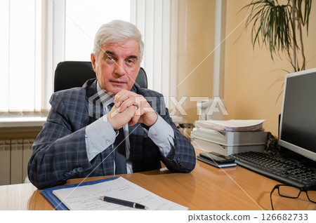 Confident elderly gray-haired man in a plaid suit sits at his desk in the office Confident elderly gray-haired man in a plaid suit sits at his desk in the office 126682733
