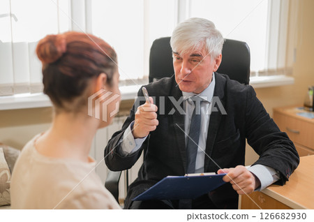 Mature gray-haired male psychotherapist with clipboard in session with female patient. Smiling caucasian woman at a psychologist appointment. Mature gray-haired male psychotherapist with clipboard in session with female patient. Smiling caucasian woman at a psychologist appointment. 126682930