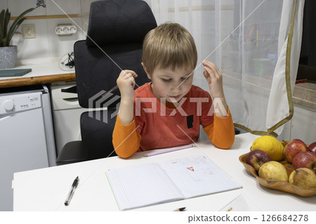Preschooler boy doing homework and holding pencil while thinking at home kitchen table 126684278