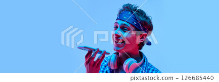 Close-up photo of young male model dressed stylish bandana and talking or recording voice message with playful smiling expression in neon light. Close-up photo of young male model dressed stylish bandana and talking or recording voice message with playful smiling expression in neon light. 126685440