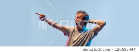 Guy pointing and yelling with big smile and eyes wide open in neon light against blue studio background showing real surprise and joy. Banner Guy pointing and yelling with big smile and eyes wide open in neon light against blue studio background showing real surprise and joy. Banner 126685447