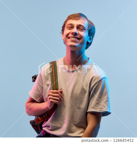Smiling boy with backpack standing in neon light against gradient background showing happy mood and relaxed feeling. Open heart and light joy Smiling boy with backpack standing in neon light against gradient background showing happy mood and relaxed feeling. Open heart and light joy 126685452