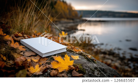 Blank white cover book resting on colorful autumn leaves by the tranquil lakeshore in fall season Blank white cover book resting on colorful autumn leaves by the tranquil lakeshore in fall season 126685507