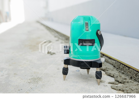 Close-up of a green laser level on a tripod, projecting straight lines to guide the placement of ceramic tiles on a white and gray floor. Close-up of a green laser level on a tripod, projecting straight lines to guide the placement of ceramic tiles on a white and gray floor. 126687966