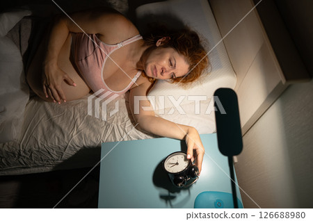 A pregnant woman lies in bed and holds an alarm clock.  126688980