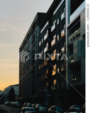 The setting sun casts a warm glow on the windows of contemporary apartment buildings, creating a serene urban scene at dusk with parked cars lining the street below. Sunset warmth illuminating windows The setting sun casts a warm glow on the windows of contemporary apartment buildings, creating a serene urban scene at dusk with parked cars lining the street below. Sunset warmth illuminating windows 126689115
