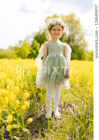 Vertical portrait of cute child girl in flower crown and fairy green dress, joyfully playing in sunny field filled with blooming yellow flowers in bright summer day. Concept of happy childhood. Vertical portrait of cute child girl in flower crown and fairy green dress, joyfully playing in sunny field filled with blooming yellow flowers in bright summer day. Concept of happy childhood. 126689937