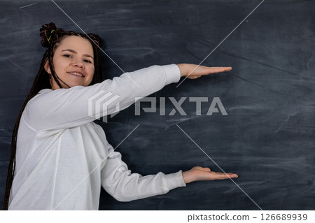 A Cheerful Girl is Gesturing Excitedly in Front of a Black Chalkboard in a Classroom Setting A Cheerful Girl is Gesturing Excitedly in Front of a Black Chalkboard in a Classroom Setting 126689939