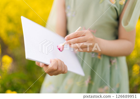 Close-up of child girl painting on blank white paper with pink paint using wooden stick standing in field of yellow flowers, creating vibrant outdoor art experience enjoying beautiful nature on summer Close-up of child girl painting on blank white paper with pink paint using wooden stick standing in field of yellow flowers, creating vibrant outdoor art experience enjoying beautiful nature on summer 126689951