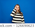 Hispanic teenage girl about 15 years old wearing striped sweater over isolated blue background gesturing thumbs up with hand smiling, looking through fingers with happy face. 126690623