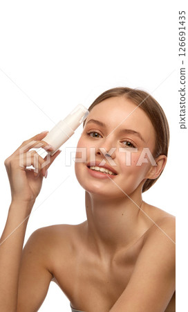 Young woman smiling while holding white cosmetic bottle near forehead, show clean healthy skin and natural makeup against white studio background. 126691435