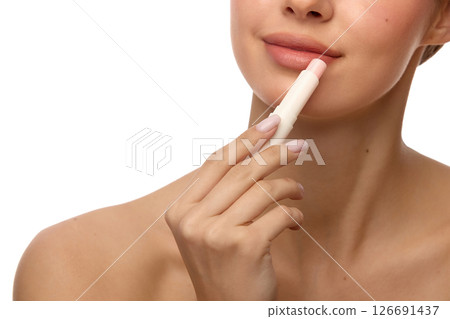 Close-up of woman applying moisturizing balm on lips with healthy skin, gentle smile and light glow on face against white background 126691437