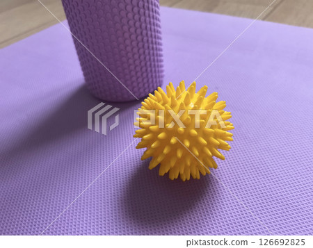 Yellow massage ball in close-up on the background of a roll for myofascial relaxation lying on a fitness and yoga mat. Sports equipment is lying on the wooden floor Yellow massage ball in close-up on the background of a roll for myofascial relaxation lying on a fitness and yoga mat. Sports equipment is lying on the wooden floor 126692825