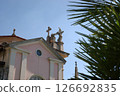 Baroque church facade with decorative crosses and statues, partially obscured by palm leaves against a clear blue sky. 126692835