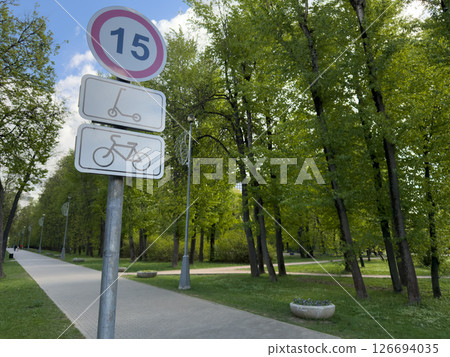 Speed limit sign for bicycles and scooters on a park pathway surrounded by tall green trees. Urban park infrastructure promoting safety and eco-friendly transportation Speed limit sign for bicycles and scooters on a park pathway surrounded by tall green trees. Urban park infrastructure promoting safety and eco-friendly transportation 126694035