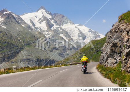 Scooter motorbike drive Grossglockner High Alpine Road mountain green hills peaks blue sky background. Winding path through highland landscape offers scenic view and mountain driving experience Scooter motorbike drive Grossglockner High Alpine Road mountain green hills peaks blue sky background. Winding path through highland landscape offers scenic view and mountain driving experience 126695687