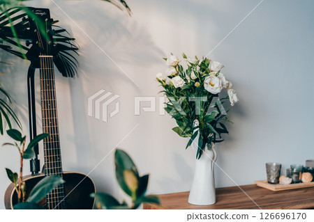 Modern minimalist style interior with modern hand-shaped vase with eustoma flowers, candles on a wooden console, guitar and home plants in sunset light on a gray wall. Cozy home interior design. Modern minimalist style interior with modern hand-shaped vase with eustoma flowers, candles on a wooden console, guitar and home plants in sunset light on a gray wall. Cozy home interior design. 126696170
