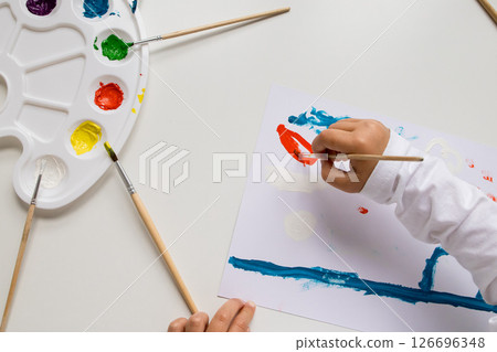 Child painting with watercolors on white paper with color palette and brushes 126696348