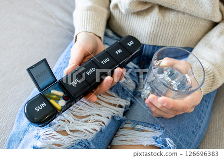 Close up of person holding pill organizer and glass of water on ripped jeans Close up of person holding pill organizer and glass of water on ripped jeans 126696383