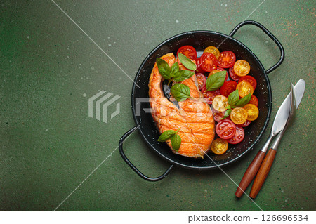 Grilled salmon steak with cherry tomatoes and basil leaves in black pan with cutlery top view on dark green background, space for text 126696534