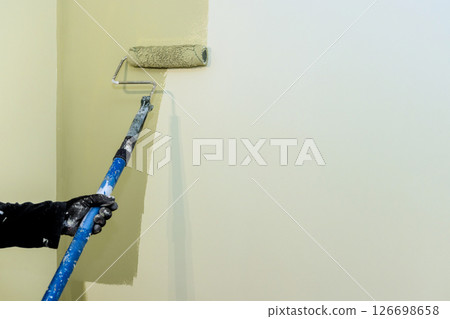 Carpenter paints wall with roller, working on home renovation project in bright living area. Carpenter paints wall with roller, working on home renovation project in bright living area. 126698658
