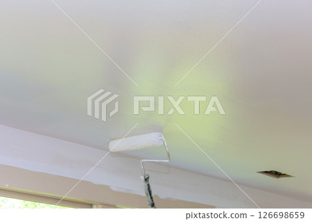 Contractor rolls paint on ceiling in spacious room filled with natural light, transforming its appearance. Contractor rolls paint on ceiling in spacious room filled with natural light, transforming its appearance. 126698659