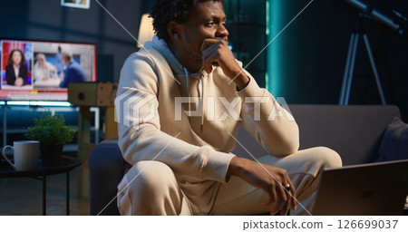 African american male remote worker sitting at home on couch late at night, overthinking about problems. Young man struggling to finish work project before deadline, finding solutions. Camera A. 126699037