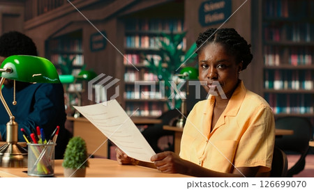 Dedicated black student reviewing papers in a traditional library setting, focusing on educational goals and study time within the campus. College girl studies and does research. Camera B. Dedicated black student reviewing papers in a traditional library setting, focusing on educational goals and study time within the campus. College girl studies and does research. Camera B. 126699070