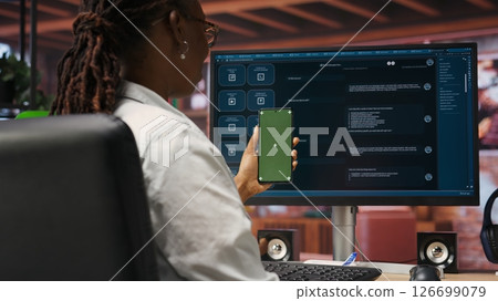 Woman uses green screen phone, doing videocall in front of PC screen with AI LLM chatbot answering prompt. Person uses large language model program during teleconference on mockup smartphone, camera B 126699079