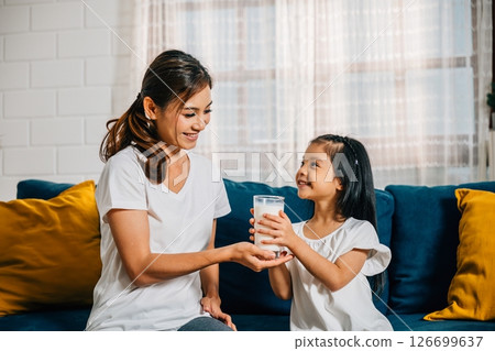 In their modern home an Asian mother lovingly offers her daughter a glass of milk on the sofa. This image reflects their carefree happiness togetherness and calcium-rich diet. 126699637