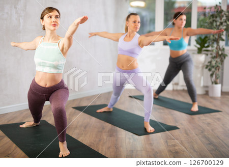 Class of focused women standing in Virabhadrasana 2 or warrior 2 position during group yoga training in gym studio Class of focused women standing in Virabhadrasana 2 or warrior 2 position during group yoga training in gym studio 126700129