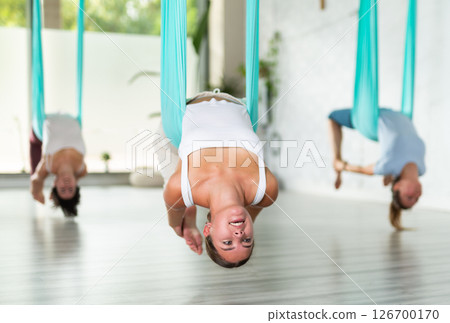 Sporty young woman using blue hammock for doing Bridge Pose Setu Bandha Sarvangasana of aerial yoga in comfortable studio with panoramic windows 126700170