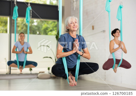 Old European female balancing in Lotus Pose Padmasana in blue hammock during fly yoga in fitness club Old European female balancing in Lotus Pose Padmasana in blue hammock during fly yoga in fitness club 126700185