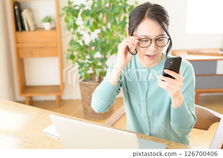 A middle-aged woman is worried while looking at a computer with a smartphone in one hand 126700406