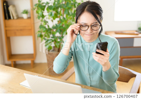 A middle-aged woman is worried while looking at a computer with a smartphone in one hand A middle-aged woman is worried while looking at a computer with a smartphone in one hand 126700407