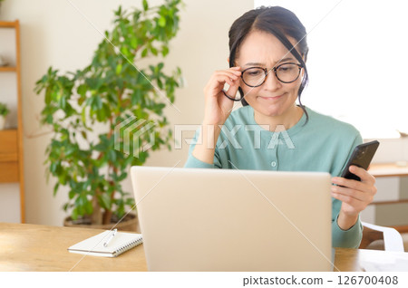A middle-aged woman is worried while looking at a computer with a smartphone in one hand A middle-aged woman is worried while looking at a computer with a smartphone in one hand 126700408