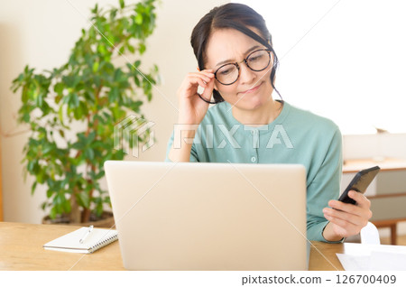 A middle-aged woman is worried while looking at a computer with a smartphone in one hand 126700409